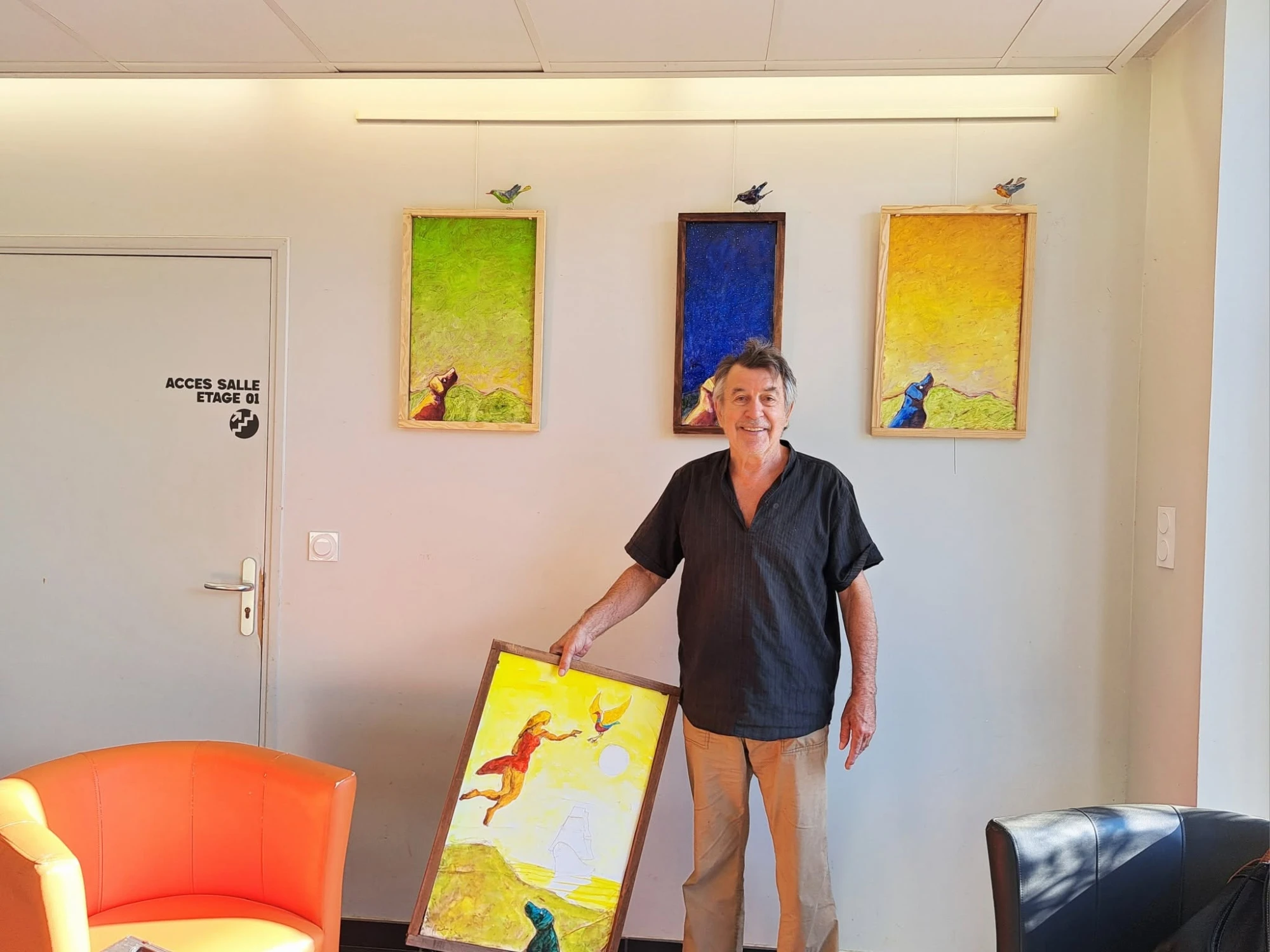 Jean-François Andre dans le hall de l’espace Louis Nodon, devant trois toiles verticales verte, bleue et jaune, tenant une toile où une femme en rouge s’élance vers un oiseau